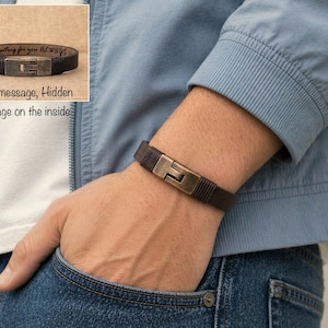 May include: A dark brown leather bracelet with a metal clasp is shown on a person's wrist. The bracelet has a rectangular clasp and a hidden message on the inside. The text on the inside of the bracelet says "these memories for you always".