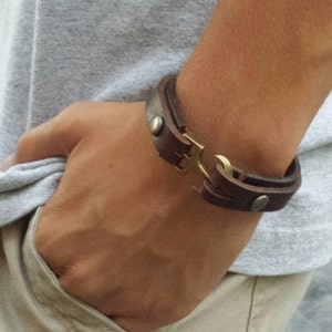 May include: A brown leather bracelet with a brass anchor clasp. The bracelet is worn on a person's wrist.