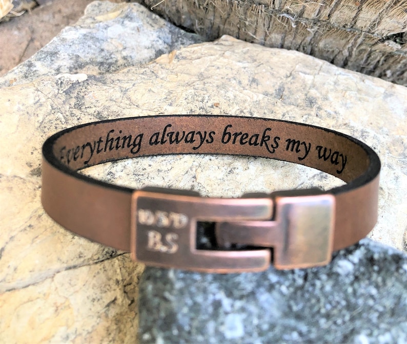 K&ouml;nnte beinhalten: Braunes Lederarmband mit einem kupfernen Verschluss. Das Armband ist mit dem Text "everything always breaks my way" graviert.