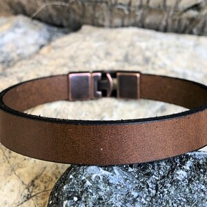 K&ouml;nnte beinhalten: Ein braunes Lederarmband mit einem kupferfarbenen Metallverschluss. Das Armband ist schlicht und elegant und eignet sich hervorragend als Geschenk f&uuml;r jeden Anlass.