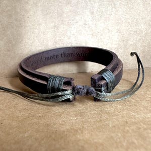 May include: A brown leather bracelet with the engraved text "loved more than words." The bracelet is secured with olive green thread and features a knotted closure. The bracelet is a simple, yet elegant accessory.