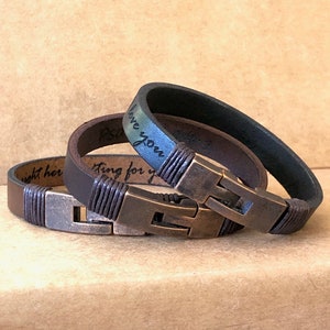 May include: Two leather bracelets with metal clasps. One is dark brown, the other is black. Both have a braided leather detail near the clasp. The bracelets have engraved text. The background is a light brown.