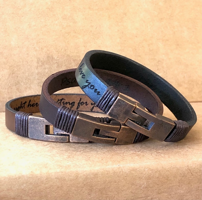 May include: Two leather bracelets with metal clasps. One is dark brown, the other is black. Both have a braided leather detail near the clasp. The bracelets have engraved text. The background is a light brown.