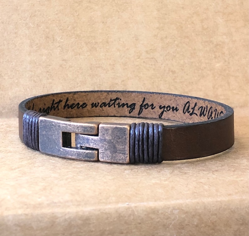 May include: Brown leather bracelet with a metal clasp. The bracelet has the engraved text: "right here waiting for you ALWAYS". The bracelet is a dark brown color.