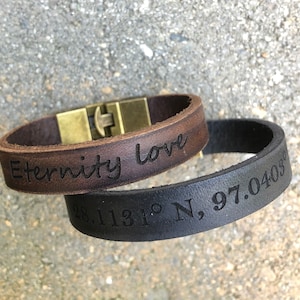 May include: Two leather bracelets with engraved text. One brown bracelet says "Eternity Love" and the other black bracelet says "28.1131° N, 97.0403°".
