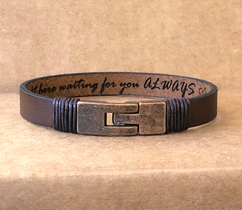 May include: A brown leather bracelet with a metal clasp. The bracelet has the words "Where waiting for you ALWAYS" engraved on the inside. The clasp is a rectangular shape with a bronze finish.