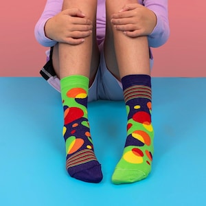 Puede incluir: Un par de calcetines coloridos con un patrón de formas abstractas en rojo, naranja, amarillo y verde sobre un fondo azul marino. Un calcetín es verde con un puño azul marino, el otro es azul marino con un puño verde.
