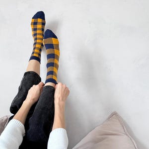May include: A pair of crew-length socks featuring a bold mustard yellow and navy blue chequered pattern. The socks have solid navy blue toes and cuffs, with a consistent grid design covering the foot and ankle area, providing a distinct contrast.