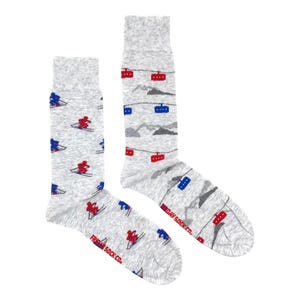 Może przedstawiać: Para szarych skarpet z wzorem w stylu narciarskim. Jedna skarpeta przedstawia narciarzy, a druga wyciągi narciarskie. Obie skarpetki mają napis "Friday Sock Co." na dole.