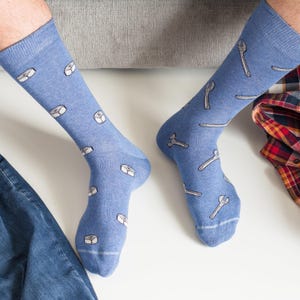 May include: A pair of light blue socks with a pattern of white nuts and bolts. The socks are worn by a person sitting down. A pair of blue jeans and a plaid shirt are also visible.
