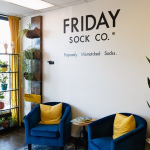 Peut inclure: Deux fauteuils bleus avec des coussins jaunes sont plac&eacute;s devant un mur blanc avec le texte "FRIDAY SOCK CO. Purposely Mismatched Socks." dessus.