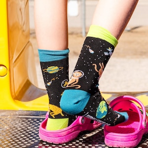 Puede incluir: Calcetines negros con temática espacial, con planetas, estrellas y un mono astronauta. Los calcetines tienen detalles en azul, verde y amarillo. Se usan con zuecos rosas brillantes.