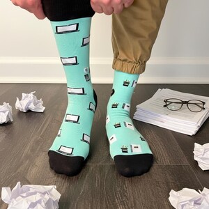 Könnte beinhalten: Socken mit hellem türkisblauem Hintergrund und schwarzen Zehen und Bündchen. Die Socken zeigen ein Muster aus weißen Computerbildschirmen, Telefonen und Büro-Symbolen. Die Person trägt eine khakifarbene Hose.