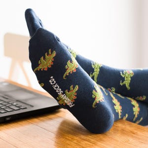Puede incluir: Un par de calcetines azules con un patrón de dinosaurio verde. Los calcetines tienen el texto "FRIDAY SOCK CO." impreso en el lateral.