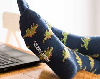 Op de afbeelding: Een paar blauwe sokken met een groen dinosauruspatroon. De sokken hebben de tekst "FRIDAY SOCK CO." op de zijkant gedrukt.