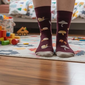 May include: Burgundy socks with a whimsical pattern of snails and mushrooms. The socks have a gray toe and heel, and the design is in shades of tan, gray, and white. The socks are on a patterned rug in a child's room.