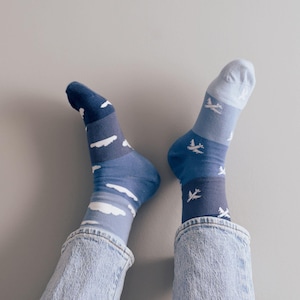 May include: A pair of blue socks with a white cloud and airplane pattern. The socks are worn by a person wearing blue jeans.