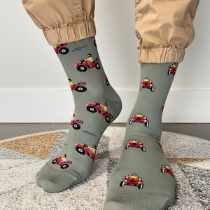 Puede incluir: Calcetines verde oliva con un estampado repetido de coches de carreras rojos y amarillos. Los calcetines los lleva una persona con pantalones color beige. Los calcetines están hechos de un material suave y cómodo.