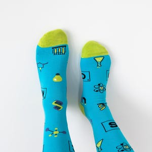 May include: A pair of blue socks with a green cuff. The socks feature a variety of science-themed illustrations, including beakers, atoms, and light bulbs.