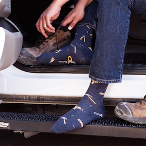 Könnte beinhalten: Ein Paar dunkelblaue Socken mit einem Muster aus gelben Werkzeugen, darunter ein Hammer, ein Schraubenschlüssel, eine Zange und ein Schraubendreher. Die Socken werden von einer Person getragen, die Jeans und braune Arbeitsschuhe trägt. Der Text "FRIDAY SOCK CO." ist auf den Socken sichtbar.