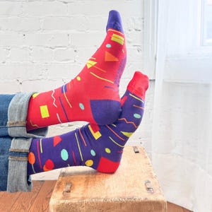 Puede incluir: Un par de calcetines con un patrón geométrico vibrante. Un calcetín es principalmente rojo con talones y puntas moradas, mientras que el otro es morado con talones y puntas rojas. Ambos están decorados con pequeñas formas amarillas, turquesas y naranjas.