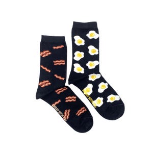 May include: A pair of black socks with a fun breakfast food design. One sock features a pattern of bacon strips, while the other sock has a pattern of fried eggs. The socks are made by Friday Sock Co.