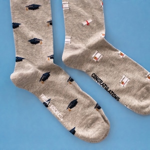 以下が含まれることがあります： 卒業帽子の模様がついたグレーの靴下と、もう一方の靴下には「おめでとうございます」と書かれたグレーの靴下のペア。靴下は「フライデースックカンパニー」製です。