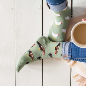 Könnte beinhalten: Ein Paar grüne Socken mit einem weißen Hühnermuster. Die Socken werden von einer Person getragen, wobei deren Jeans sichtbar sind.