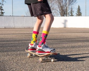 Puede incluir: Una persona en una patineta con calcetines y zapatillas coloridas. Los calcetines tienen un estampado de dinosaurios de dibujos animados sobre un fondo amarillo y rosa. Las zapatillas son blancas, rojas y grises. La patineta es marrón con ruedas blancas.