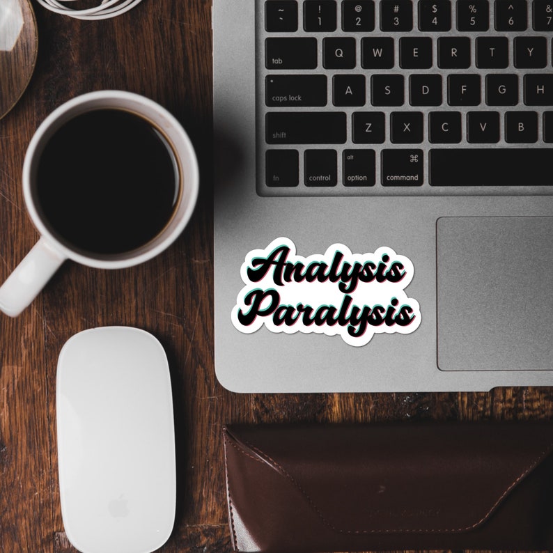 May include: A white sticker with the words "Analysis Paralysis" in a black, green, and white 3D effect font, placed on a laptop. The laptop is on a wooden surface, with a coffee cup and a computer mouse.