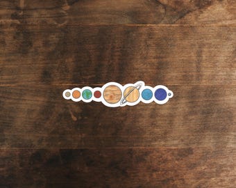 May include: A sticker featuring a colorful illustration of the solar system's planets in a row. The planets are depicted in their respective colors, with Saturn's rings clearly visible. The sticker is on a wooden surface.