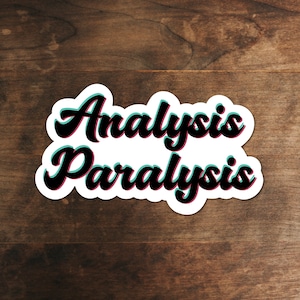 May include: A white sticker with the words "Analysis Paralysis" in a black, cursive font, outlined in teal and pink. The sticker is placed on a dark brown, wooden surface.