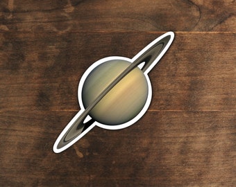 May include: A sticker of Saturn, a gas giant planet with a ring system, on a brown wooden background.