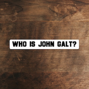 May include: White sticker with black text that reads "WHO IS JOHN GALT?" on a brown wooden background.
