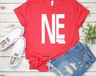 May include: A red heather t-shirt with a white distressed graphic of the letters "NE" on it. The shirt is laid flat on a white wooden surface with a pair of white sneakers and a pair of blue denim shorts.