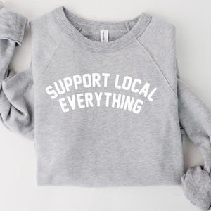May include: A heather gray sweatshirt with the text "SUPPORT LOCAL EVERYTHING" in white block letters. The raglan sleeves are folded, and the crew neck is visible. The sweatshirt is made of a soft material.