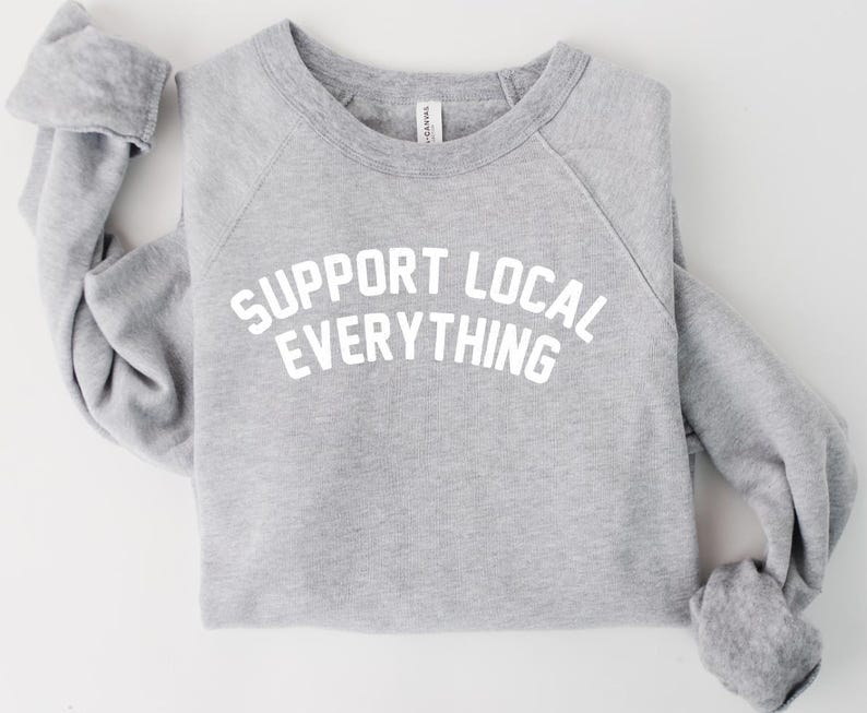 May include: A heather gray sweatshirt with the text "SUPPORT LOCAL EVERYTHING" in white block letters. The raglan sleeves are folded, and the crew neck is visible. The sweatshirt is made of a soft material.