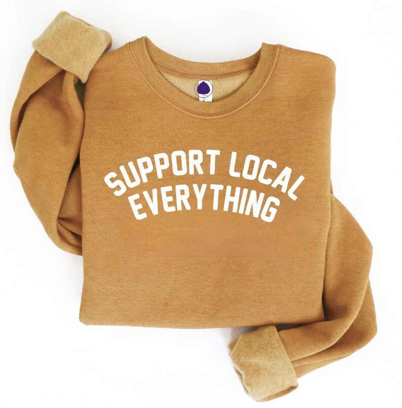 May include: A tan-colored sweatshirt with the words "SUPPORT LOCAL EVERYTHING" printed in white across the front. The sweatshirt has a crew neck and long sleeves with folded cuffs. The background is white.