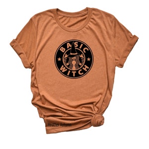 May include: A brown t-shirt with a black graphic of a witch wearing a hat and the text "BASIC WITCH" in a circle.