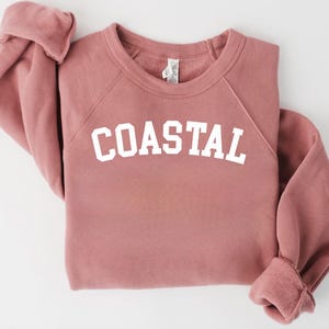 May include: A dusty rose-colored sweatshirt with the word "COASTAL" printed in bold white letters across the chest. The sweatshirt has a crew neck and raglan sleeves. The cuffs and hem are ribbed.