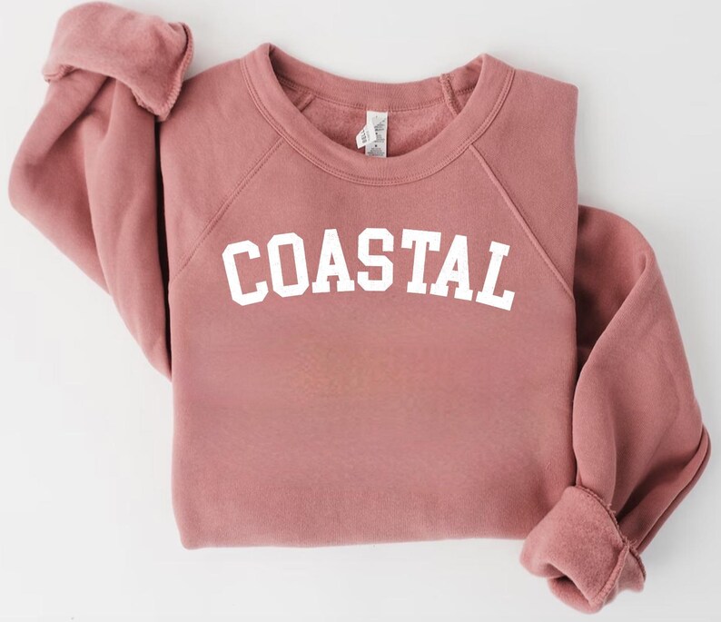 May include: A dusty rose-colored sweatshirt with the word "COASTAL" printed in bold white letters across the chest. The sweatshirt has a crew neck and raglan sleeves. The cuffs and hem are ribbed.