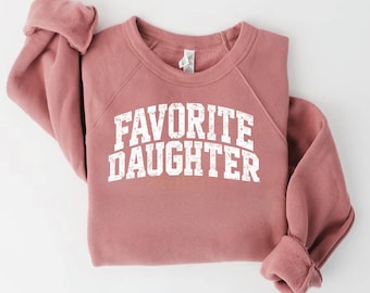 Op de afbeelding: Een oudroze sweater met ronde hals en de tekst FAVORITE DAUGHTER in een wit, verweerd lettertype in universiteitsstijl op de borst. Het kledingstuk heeft lange mouwen met geribbelde boorden en een geribbelde halslijn.