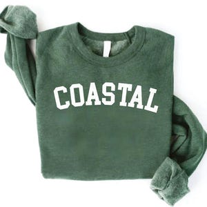 May include: A dark green sweatshirt with the word "COASTAL" printed in large, white, block letters across the chest. The sweatshirt has a crew neck and long sleeves, with the sleeves folded up. The background is white.