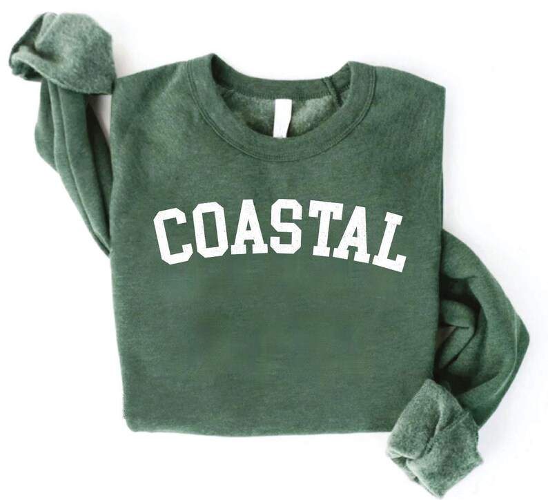 May include: A dark green sweatshirt with the word "COASTAL" printed in large, white, block letters across the chest. The sweatshirt has a crew neck and long sleeves, with the sleeves folded up. The background is white.