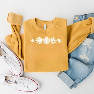 May include: A mustard yellow sweatshirt with a white floral design on the chest. The sweatshirt is paired with blue jeans and white sneakers.