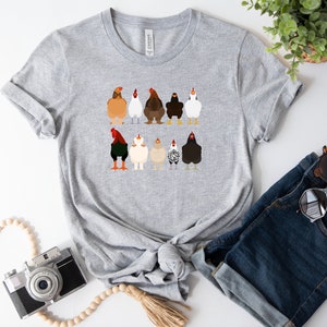 May include: A grey t-shirt featuring a graphic of nine chickens in various colours and patterns. The chickens are facing forwards with their tails raised.