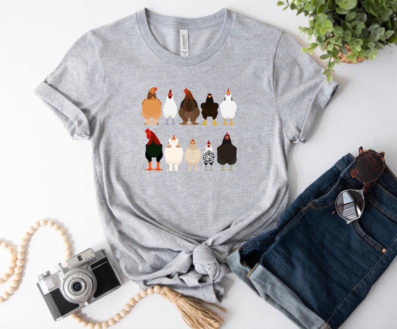 May include: A grey t-shirt featuring a graphic of nine chickens in various colours and patterns. The chickens are facing forwards with their tails raised.