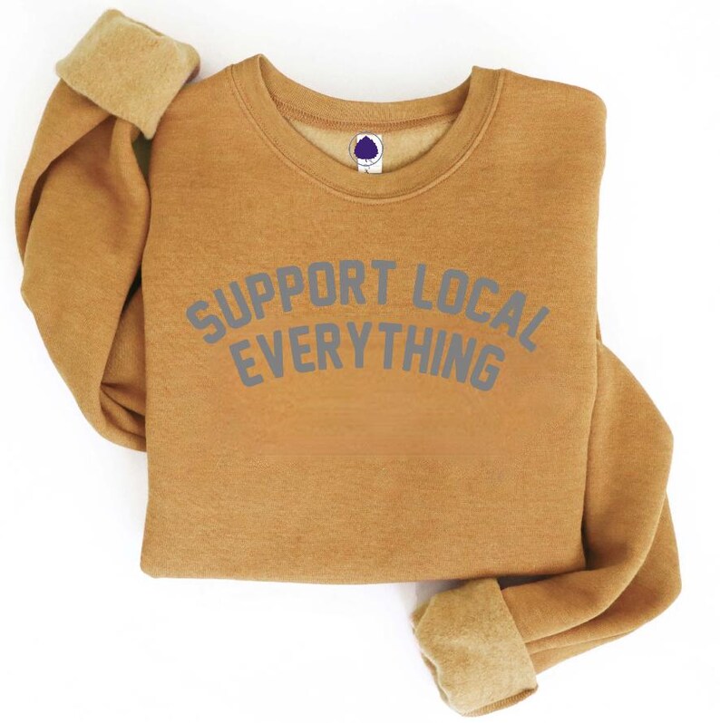 May include: Mustard-colored sweatshirt with the text "SUPPORT LOCAL EVERYTHING" in gray lettering. The sweatshirt has a crew neck and ribbed cuffs. The sleeves are folded up, revealing a lighter tan lining. The garment is made of a soft material.