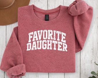 Op de afbeelding: Een rood gemêleerd sweatshirt met ronde hals en de tekst FAVORITE DAUGHTER in een verweerd wit collegiaal lettertype op de borst gedrukt. Het kledingstuk heeft lange mouwen en een zwart label aan de binnenkant van de kraag.