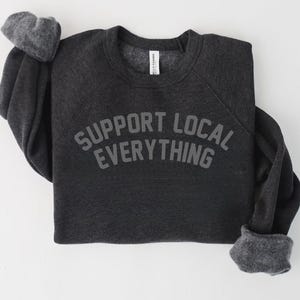 May include: Dark gray sweatshirt with the text "SUPPORT LOCAL EVERYTHING" in gray block letters. The sweatshirt has long sleeves with gray cuffs and a crew neck. The garment is made of a soft material.